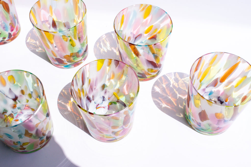 Six hand-blown glass tumblers with colorful, marbled patterns of pink, yellow, green, and orange on a bright white background, casting strong shadows.